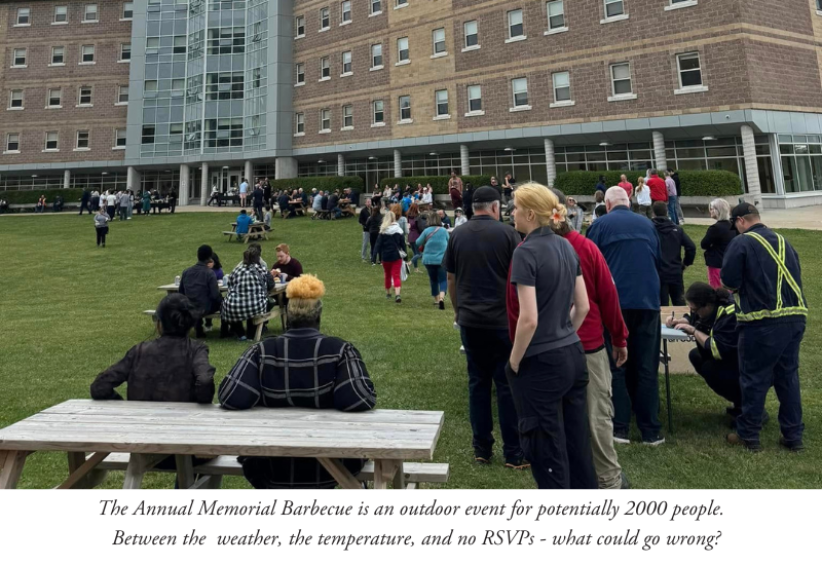 Image of people lining up for a barbecue, picnic tables and a building in the background